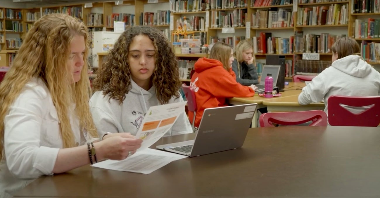 Two persons look at a computer and worksheet